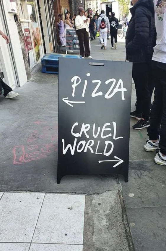 Signboard on a sidewalk with arrows pointing left to "PIZZA" and right to "CRUEL WORLD."