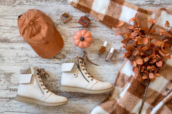 Flat lay of a laid-back autumn outfit featuring a plaid fleece jacket, white boots, a baseball cap, nail polishes, eyeshadows, a faux pumpkin, and leaves