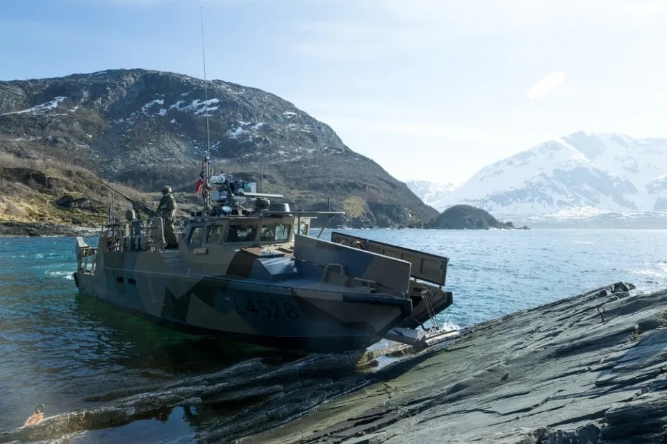 US Marines on a CB90 boat that has reached a rocky shore, with snowy mountains in the background.