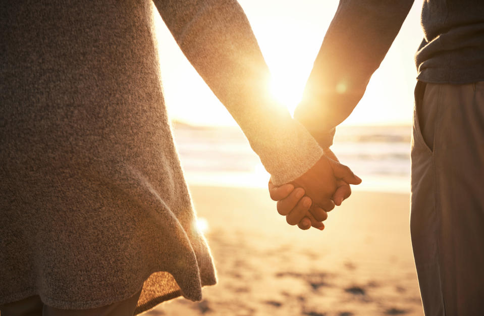 Two people holding hands on a sunlit beach. Their faces are not visible, focusing on their hands and the warm, serene setting