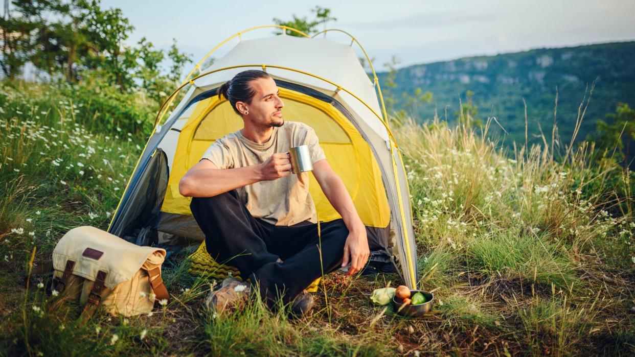  Man sitting on the grass outside his tent having a hot drink. 