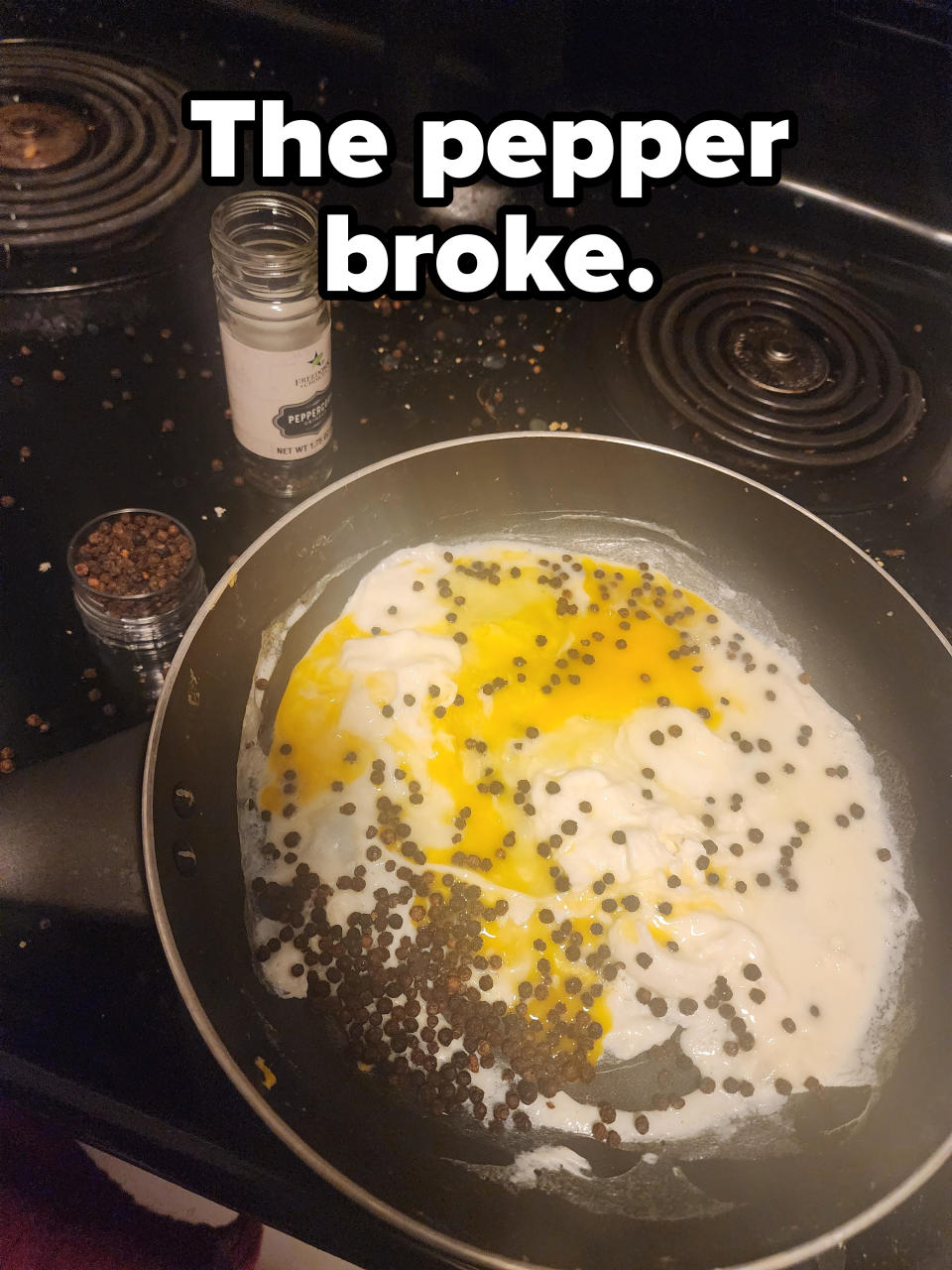 A pan on a stove with eggs cooking, heavily seasoned with whole black peppercorns. An open pepper bottle and scattered peppercorns are on the stove