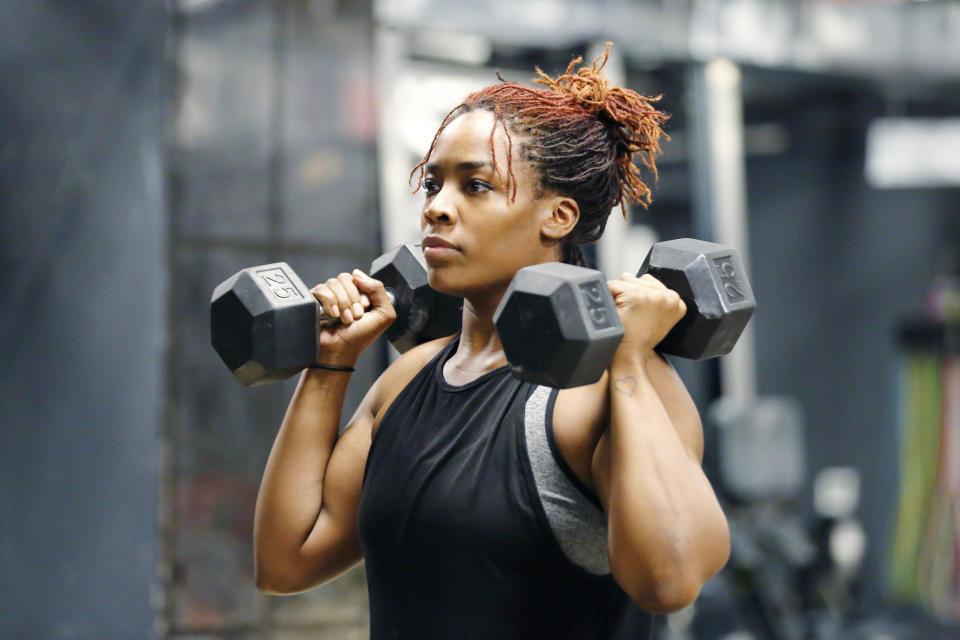 A woman lifts two dumbbells on her shoulders during a workout session at a gym