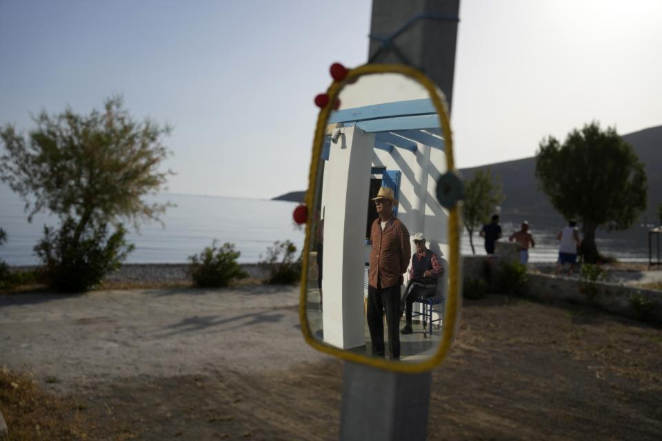 Hotel owner Akis Sartzetakis and his friend are reflected in a mirror as three men jog past next to the beach on the Aegean Sea island of Tilos, southeastern Greece, Tuesday, May 10, 2022. When deciding where to test green tech, Greek policymakers picked the remotest point on the map, tiny Tilos. Providing electricity and basic services, and even access by ferry is all a challenge for this island of just 500 year-round inhabitants. It's latest mission: Dealing with plastic. (AP Photo/Thanassis Stavrakis)