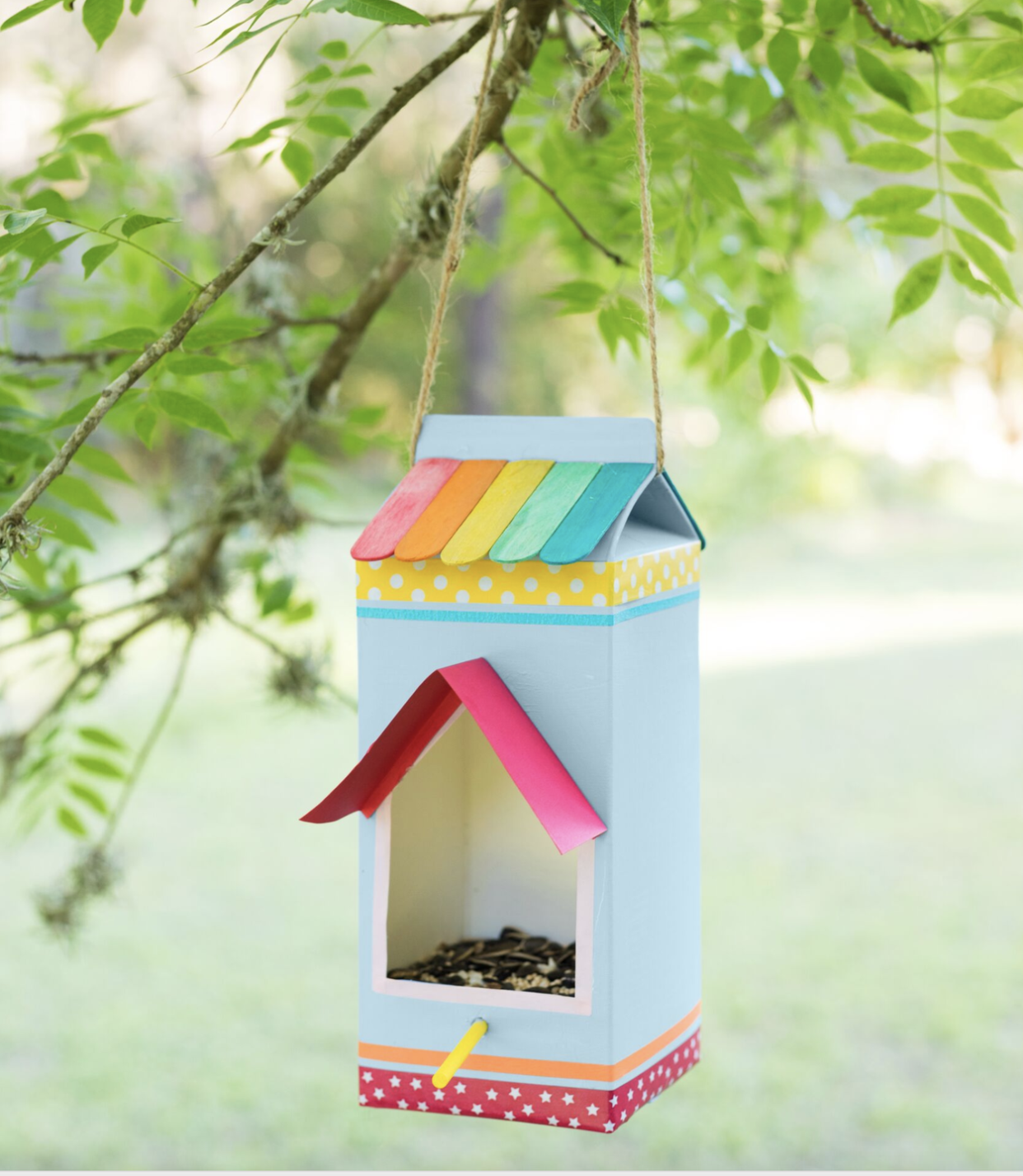 a milk carton decorated with paper washi tape and colorful popsicle sticks turned into a bird feeder hanging from a branch