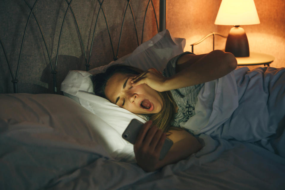 Girl lying in bed uses a cell phone and yawns. She is tired or insomnia or addicted to the phone