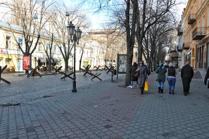 Anti-tank obstacles line an urban road as pedestrians walk along the sidewalk.