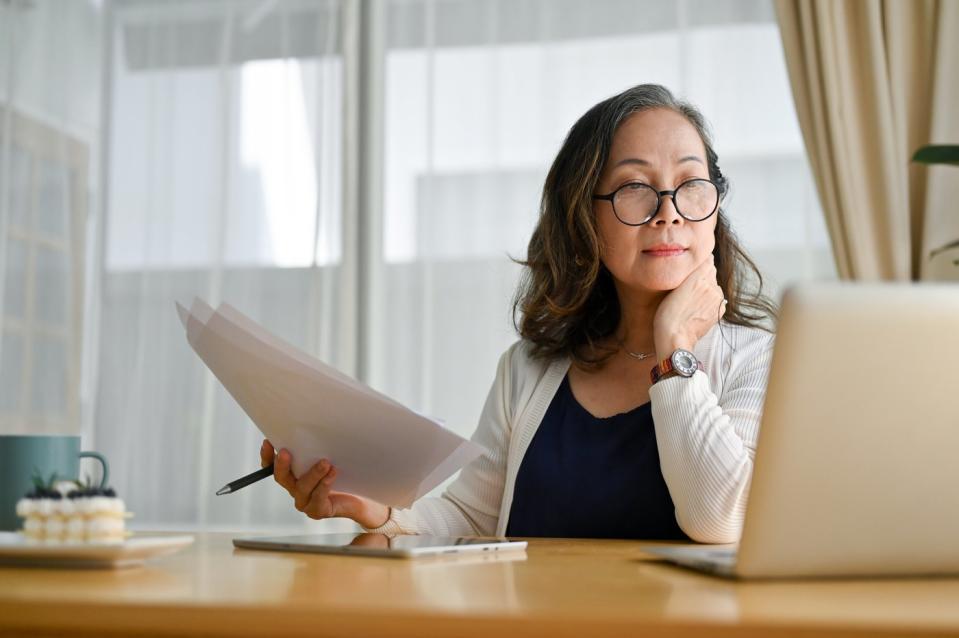 A person sitting in an office who's holding paperwork in their right hand and looking at an open laptop on a desk.