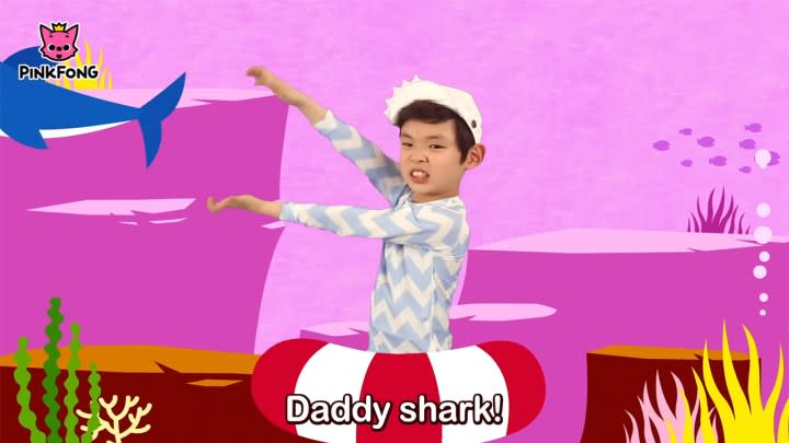 A child stretching and moving his arms like a shark's mouth with animation behind him in the Pinking Baby Shark video on YouTube.