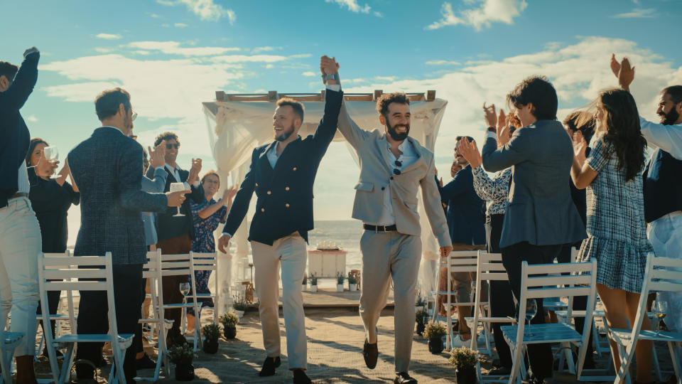 Two men in suits raise their hands in celebration as they walk down an aisle at an outdoor event surrounded by clapping guests