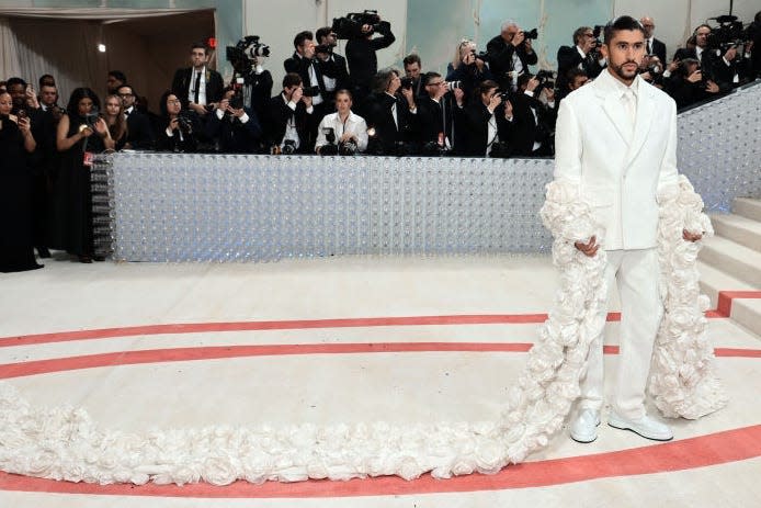 Bad Bunny attends the 2023 Met Gala wearing a white suit with a floral train draped on his arms.