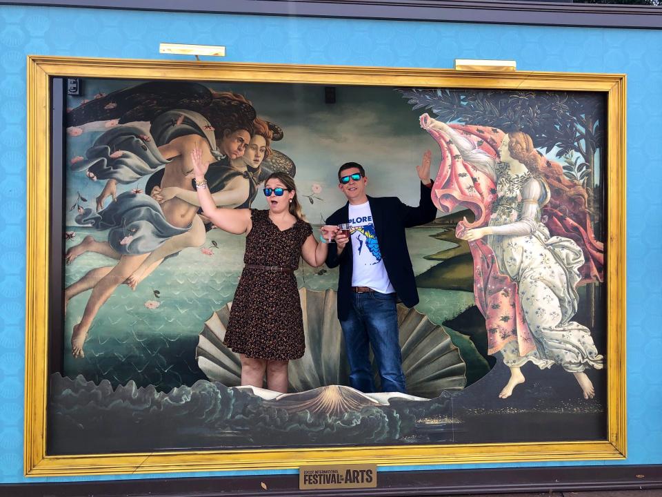 Terri Peters and her husband posing in a frame depicting the painting The Birth of Venus. They stand where Venus should be in front of a shell. Terri has dyed blonde hair and wears blue reflective sunglasses and a black dress with a floral pattern. Her husband wears the same sunglasses, a black blazer, white t-shirt with graphic design, and blue jeans. In front of the painting is the sign that says "Festival of the Arts."