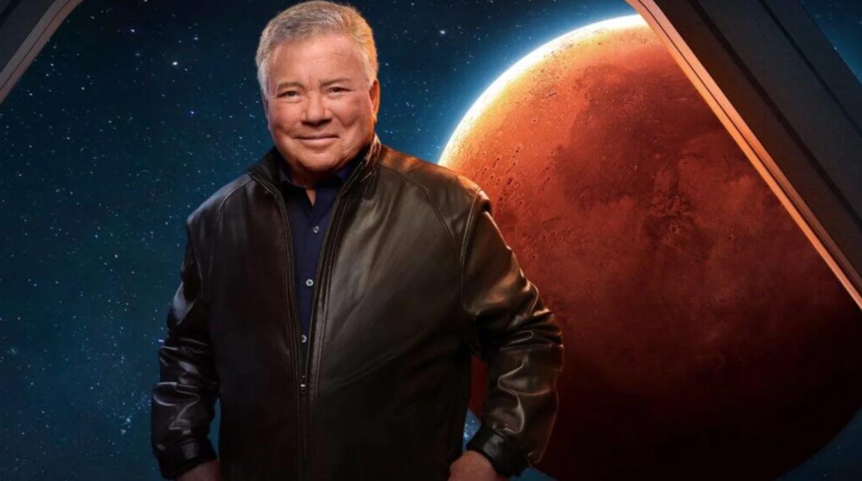  william shatner stands in front of simulated spaceship window, which shows mars in the background. 