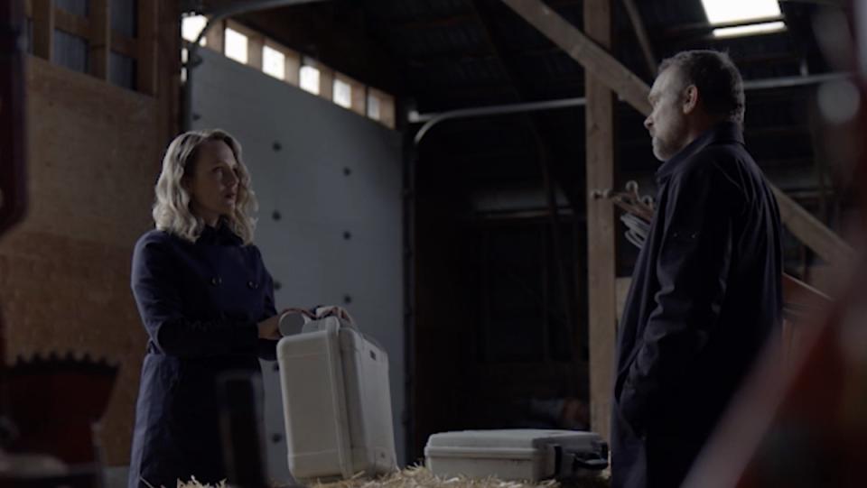 A blonde woman holds a white case as a man with a case in front of him on hay in a barn looks at her