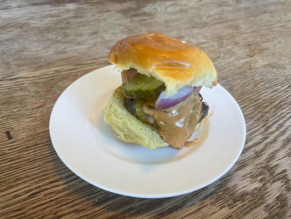 A slider with melted cheese, an orange sauce, sliced pickle, and onion between a bun on a white plate