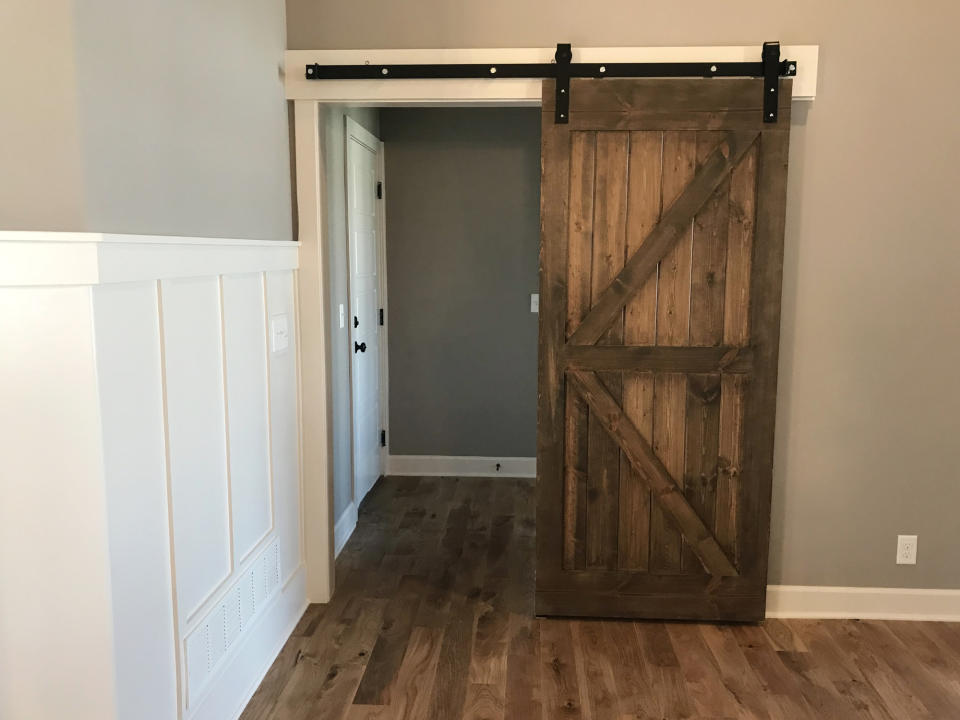 A wooden barn door on a black sliding track is partially open, revealing a hallway. The room has hardwood floors and white wainscoting on one wall