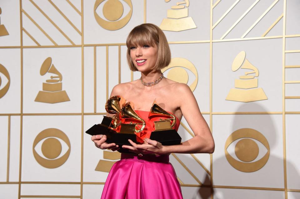 taylor swift smiles and stands while holding three grammy awards, she wears a strapless orange and hot pink gown with a gold chocker necklace