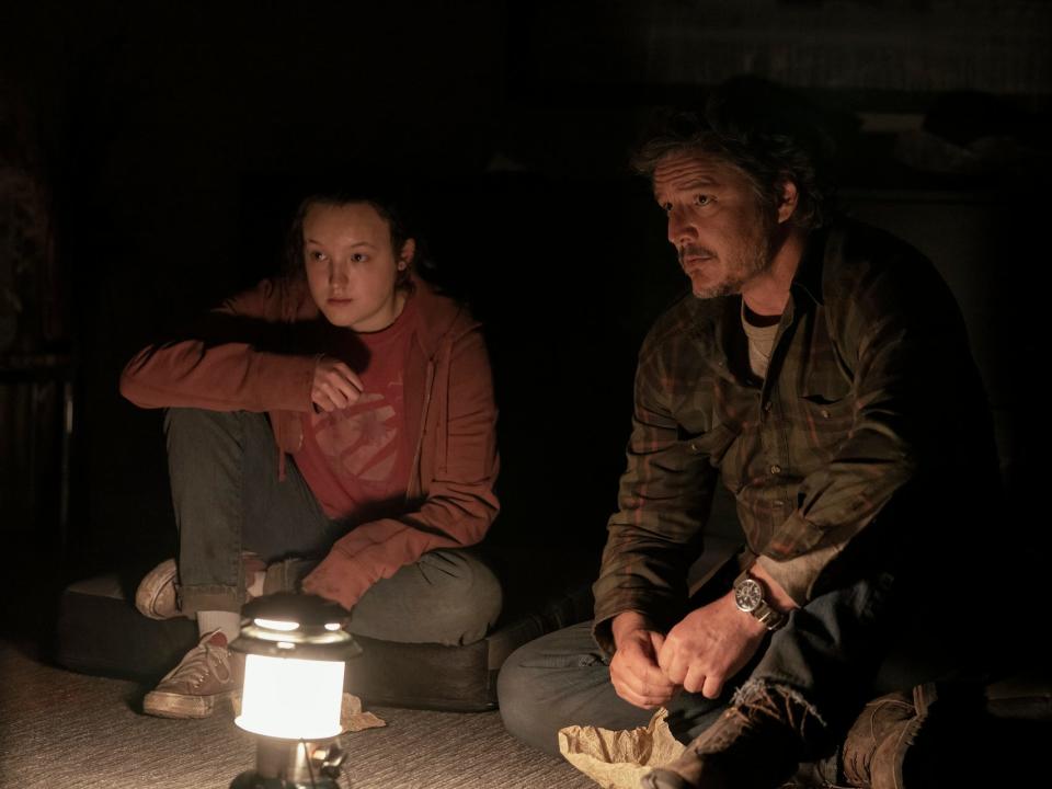 bella ramsey and pedro pascal in the last of us as ellie and joel. they're sitting on a carpeted floor, in front of a small camp light.