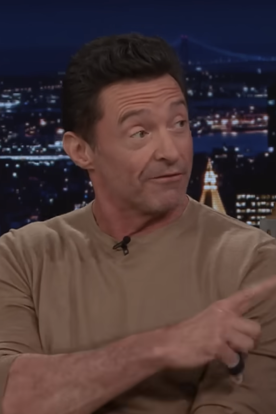 Hugh Jackman is gesturing while sitting on a talk show set, with a nighttime cityscape in the background