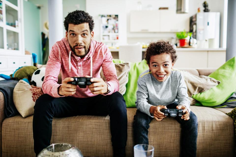 A parent and child sit on a couch and hold controllers while playing video games.