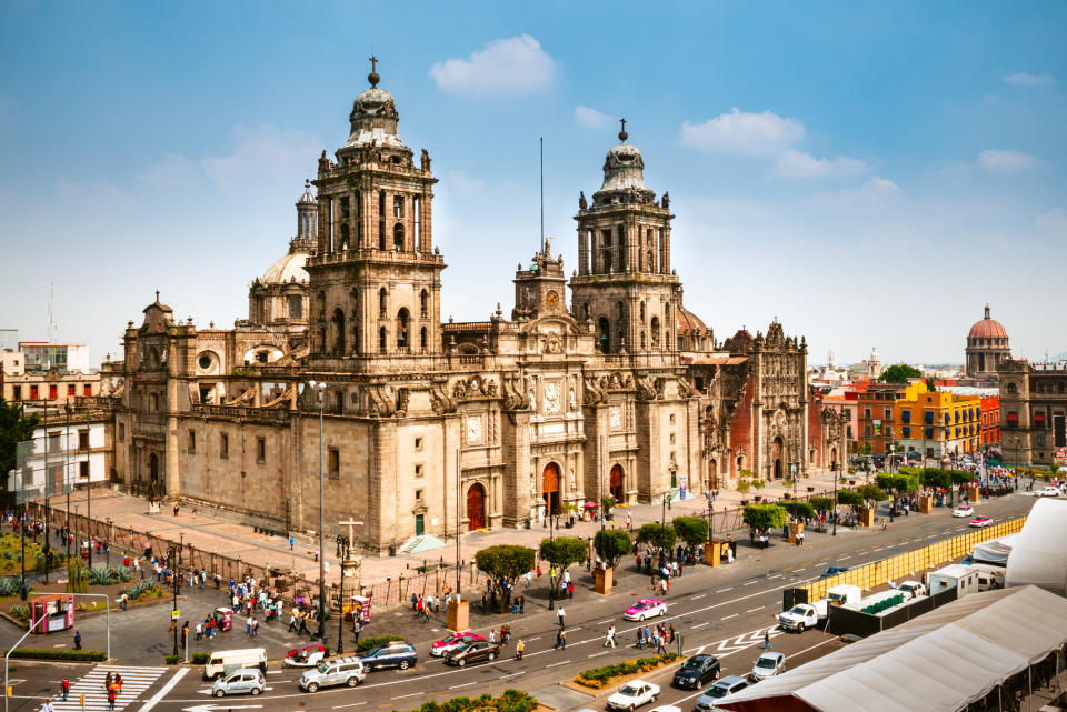People walking and cars driving in front of the Metropolitan Cathedral in Mexico City, with surrounding buildings visible in the background
