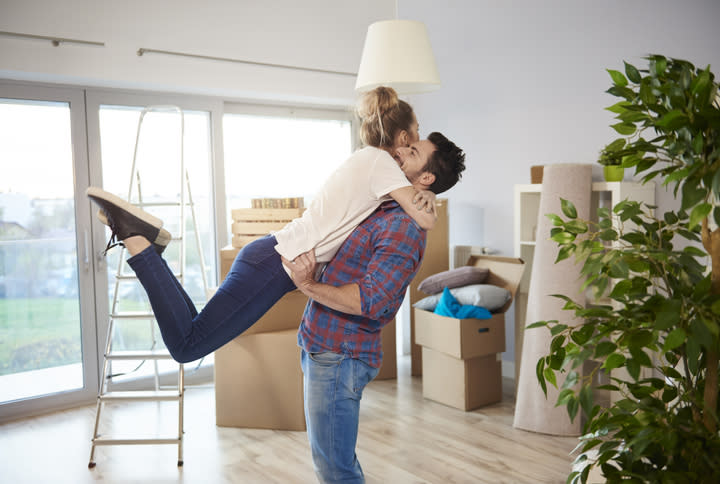Young couple at home, surrounded by cardboard boxes, man lifting woman in excited hug.