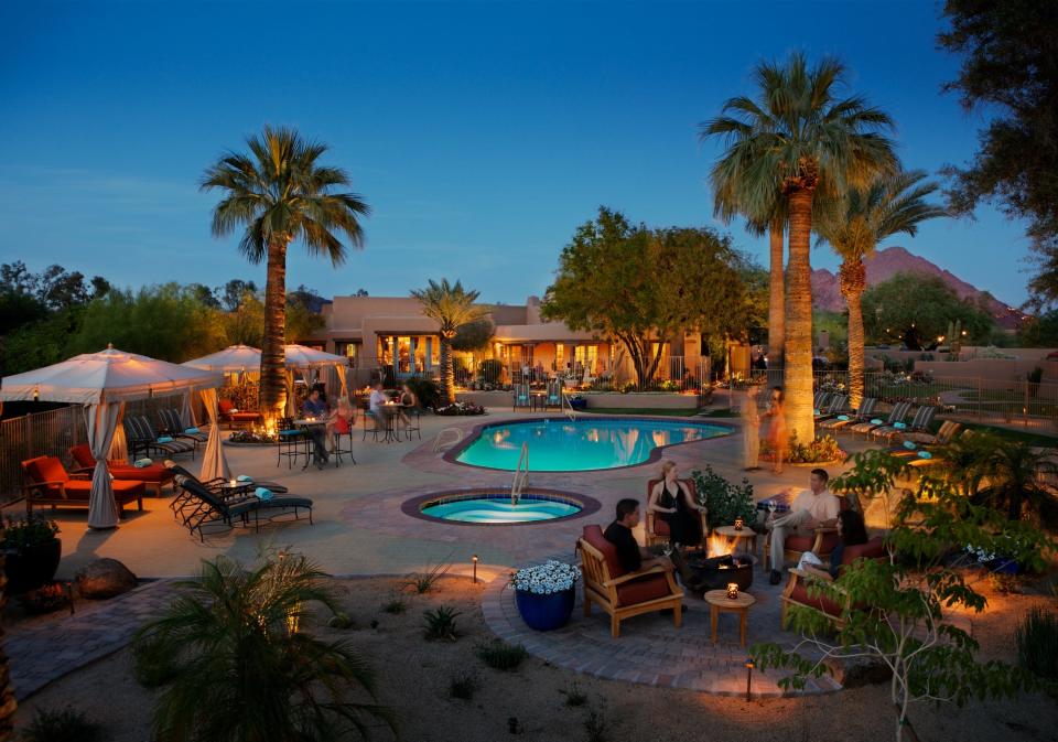 A pool lit up with palm trees around it and an adobe hotel behind it. Mountains in the background at dusk