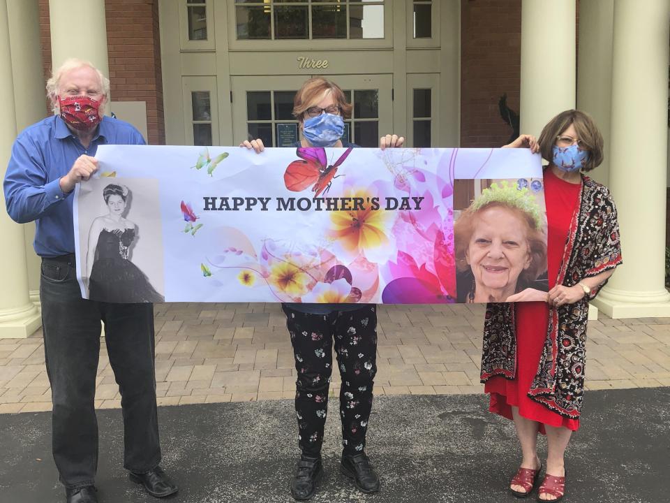 This May 3, 2020, photo released by Shelly Solomon shows, from left, Steve Turner and his sisters, Carla Paull and Lisa Fishman, holding up a Mother’s Day banner emblazoned with images of their mom, Beverly Turner, in front of her assisted living facility in Ladue, Missouri. They were “practicing” how their Mother’s Day surprise will look on the holiday as their mother peers down from a window. Isolation due to the coronavirus outbreak has led mothers and offspring to find creative ways to celebrate. (Shelly Solomon via AP)