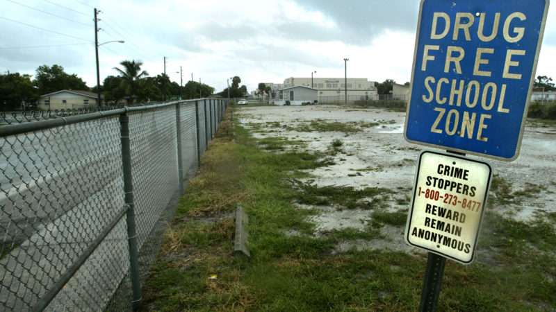 A 'drug free school zone' sign stands on a post next to a fence