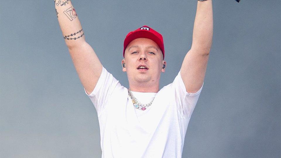 Aitch, a male rapper on stage, wearing a red cap, white tshirt with a necklace - with his arms raised high mid performance.