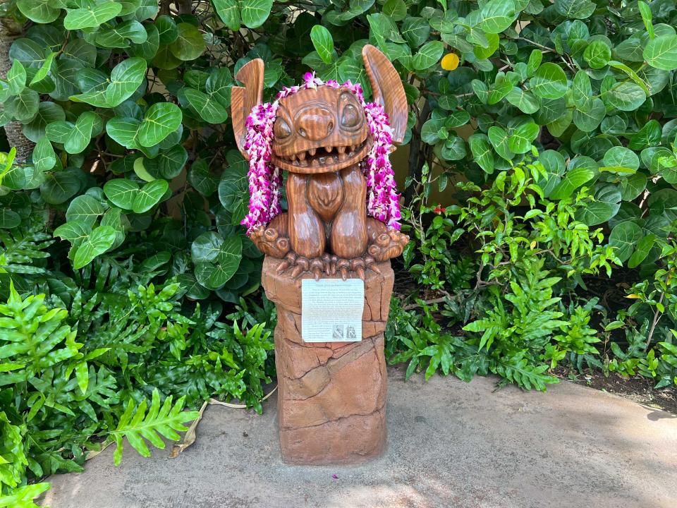 A wooden statue of Stitchy from "Lilo and Stitch" draped in flower leis with a sign underneath in front of green bushes.
