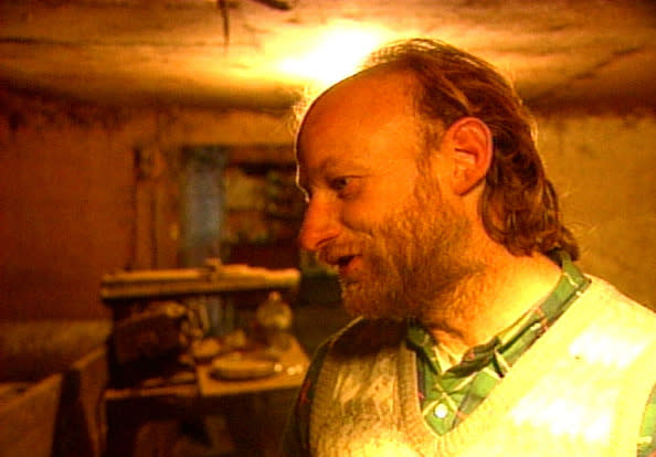 A man with a beard and long hair is in a dimly lit room filled with various tools and objects. He is wearing a plaid shirt and a sweater