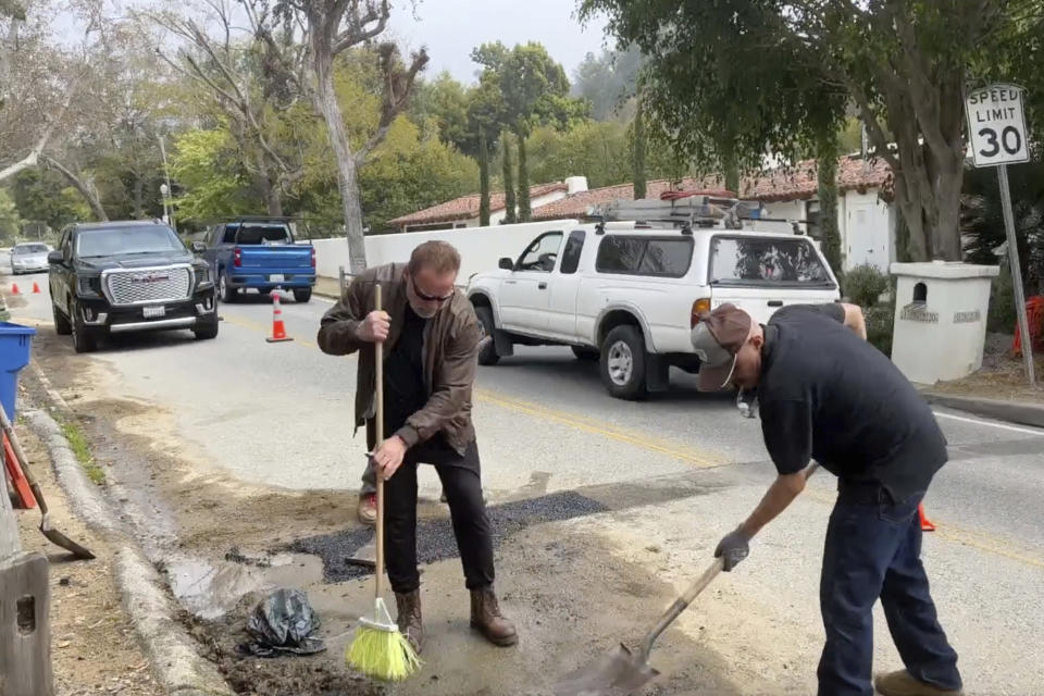 This video still image provided by The Office of Arnold Schwarzenegger, shows former California Governor Arnold Schwarzenegger, left, repairing a pot hole on a street in his Los Angeles neighborhood on Tuesday, April 11, 2023. Fed up by an enormous pothole in his neighborhood, Schwarzenegger picked up a shovel and filled it himself. (The Office of Arnold Schwarzenegger via AP)