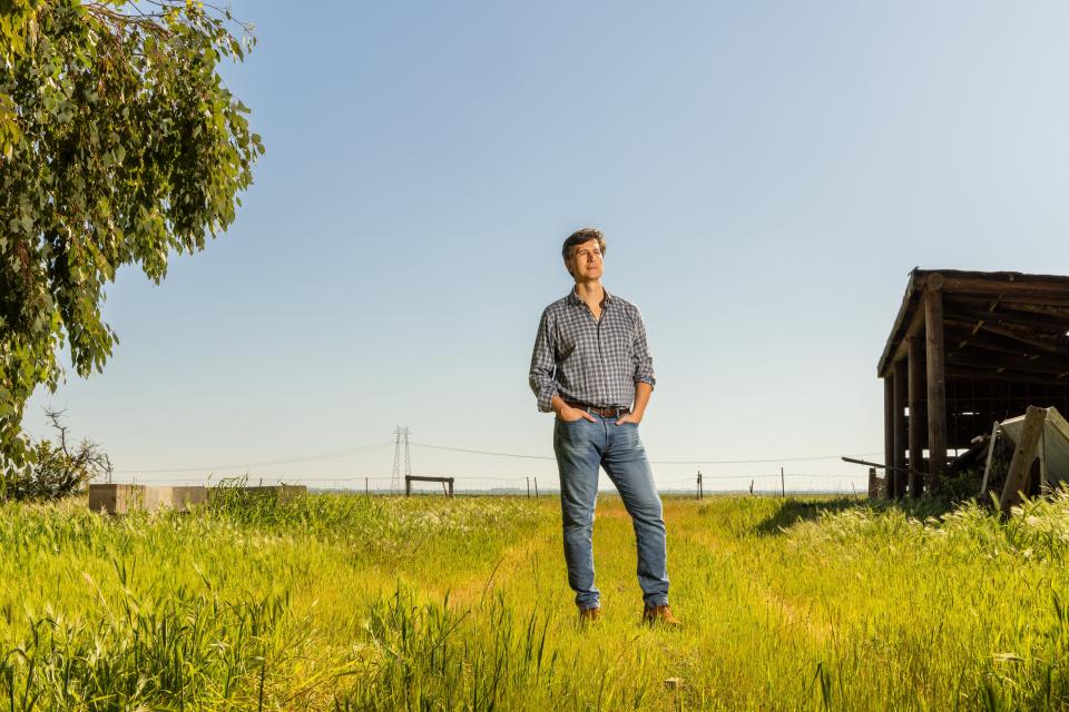 Jan Sramek standing in wide field of grass with powerlines in the and a wooden shed in the back