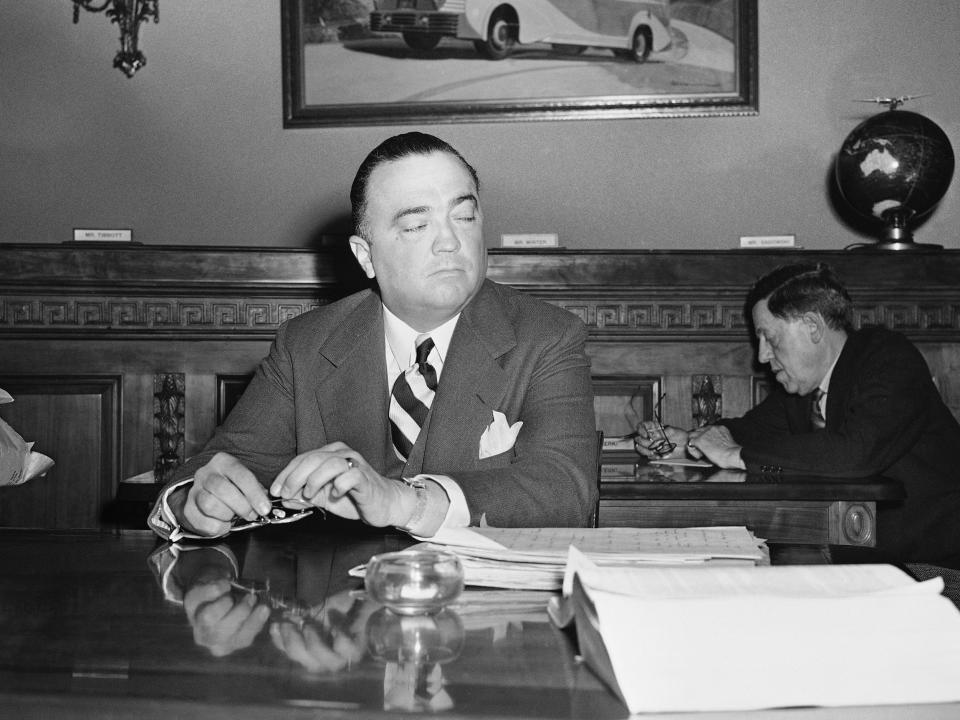 J. Edgar Hoover sits at a desk with his eyes closed.