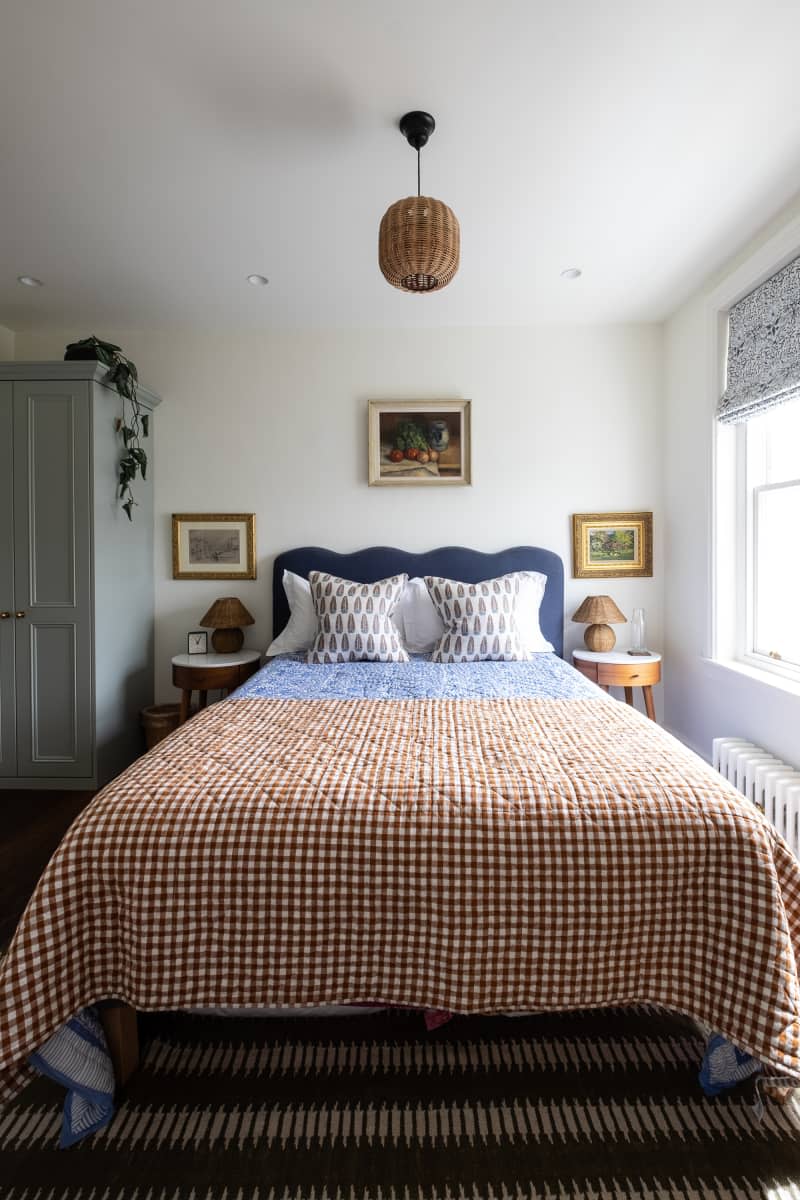 Blue upholstered scalloped headboard in bedroom with neatly made bed with brown and white gingham quilt. Small portraits hung around bed with   straw pendant hung above bed.
