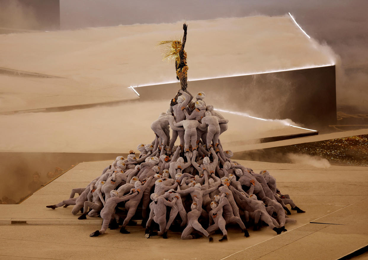 The Golden Voyager stands at the apex of an assemblage of masked dancers.