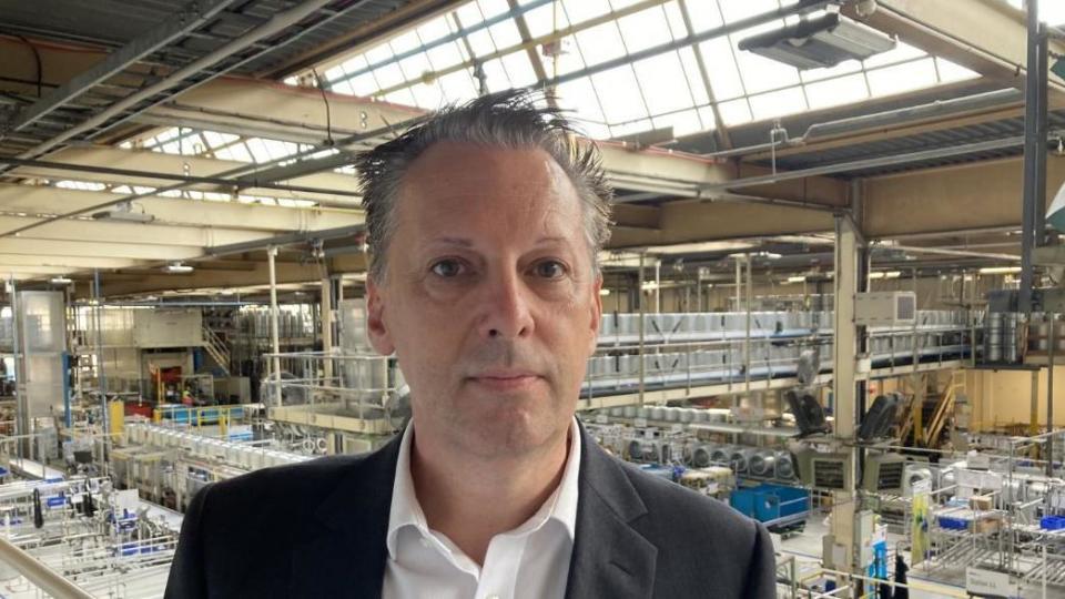 A man in a grey suit standing in factory with workers in the background