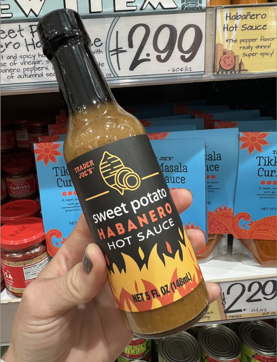 A hand holds a bottle of Trader Joe's Sweet Potato Habanero Hot Sauce in front of store shelves displaying other sauces. The price tag reads $2.99