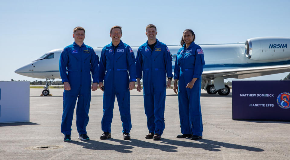 Four astronauts in their blue NASA flight suits smile as they stand on a plane tarmac with an aircraft behind them