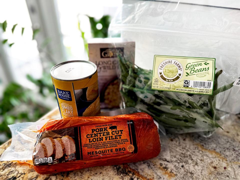 A package of pork loin with an orange marinade, a clear plastic bag of green beans, a gray box of rice, and a yellow can of biscuits on a stone countertop