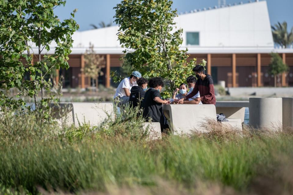 A group of teens wearing masks sit at picnic table surrounded by greenery