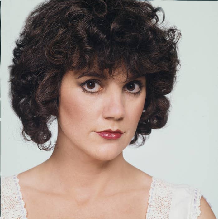 Woman with curly short hair poses with serious expression wearing white top with lace trim. No other identifiable persons present