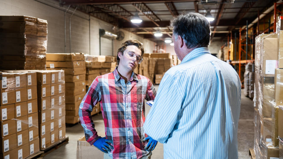 A mature warehouse manager discusses issues with a younger male warehouse worker.