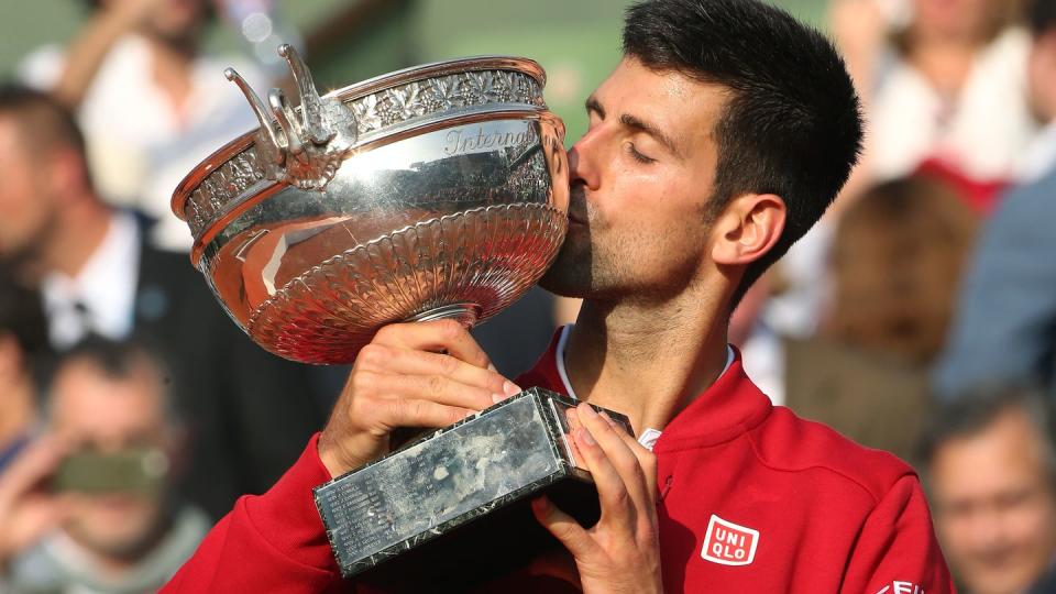 novak djokovic holds a silver trophy as he kisses it, he wears a red jacket and watch