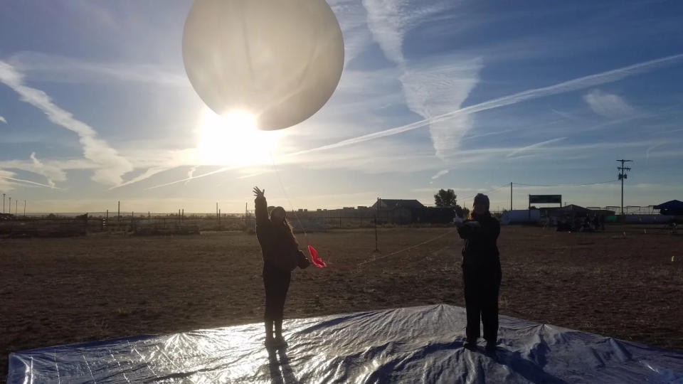     Zwei Menschen stehen auf einer Plane auf einer Wiese und lassen einen großen weißen Ballon in die Luft steigen. Die Sonne scheint dahinter, während Kondensstreifen den blauen Himmel darüber ziehen. 