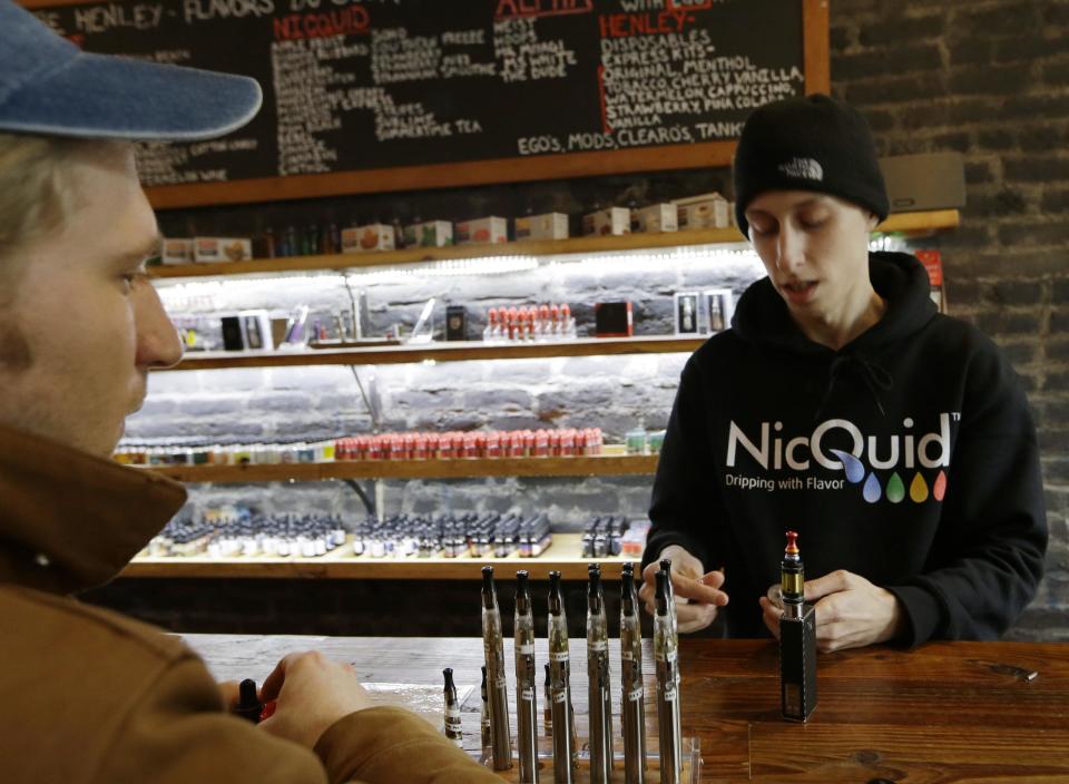 In this Feb. 20, 2014 photo, customer Patrick Johnson, left, watches as Nicholas Vukan discusses e-cigarettes at the Henley Vaporium in New York. E-cigarettes are usually made of metal parts combined with plastic or glass and come in a variety of shapes and sizes. They heat the liquid nicotine solution, creating vapor that quickly dissipates when exhaled. The vapor looks like tobacco smoke and can feel like tobacco smoke when taken into the lungs at varying strengths, from no nicotine up to 24 milligrams or more. (AP Photo/Frank Franklin II)