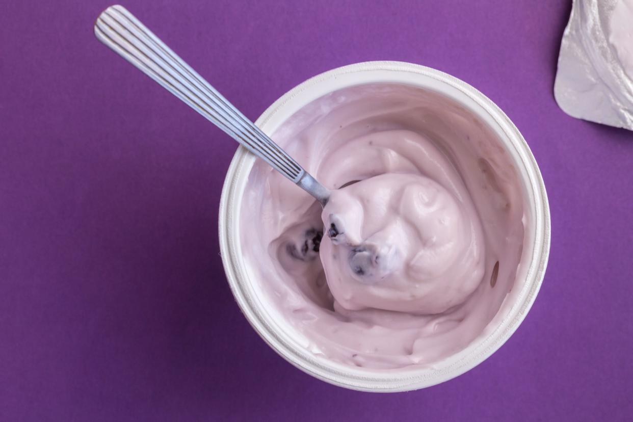 Top view of a yogurt cup with blueberry yogurt with spoon, foil top on the side, against a purple background