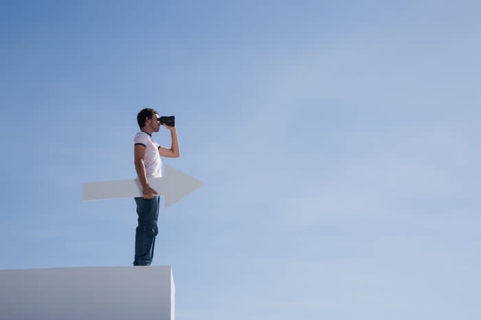 A person stands on a roof holding an arrow and a pair of binoculars.