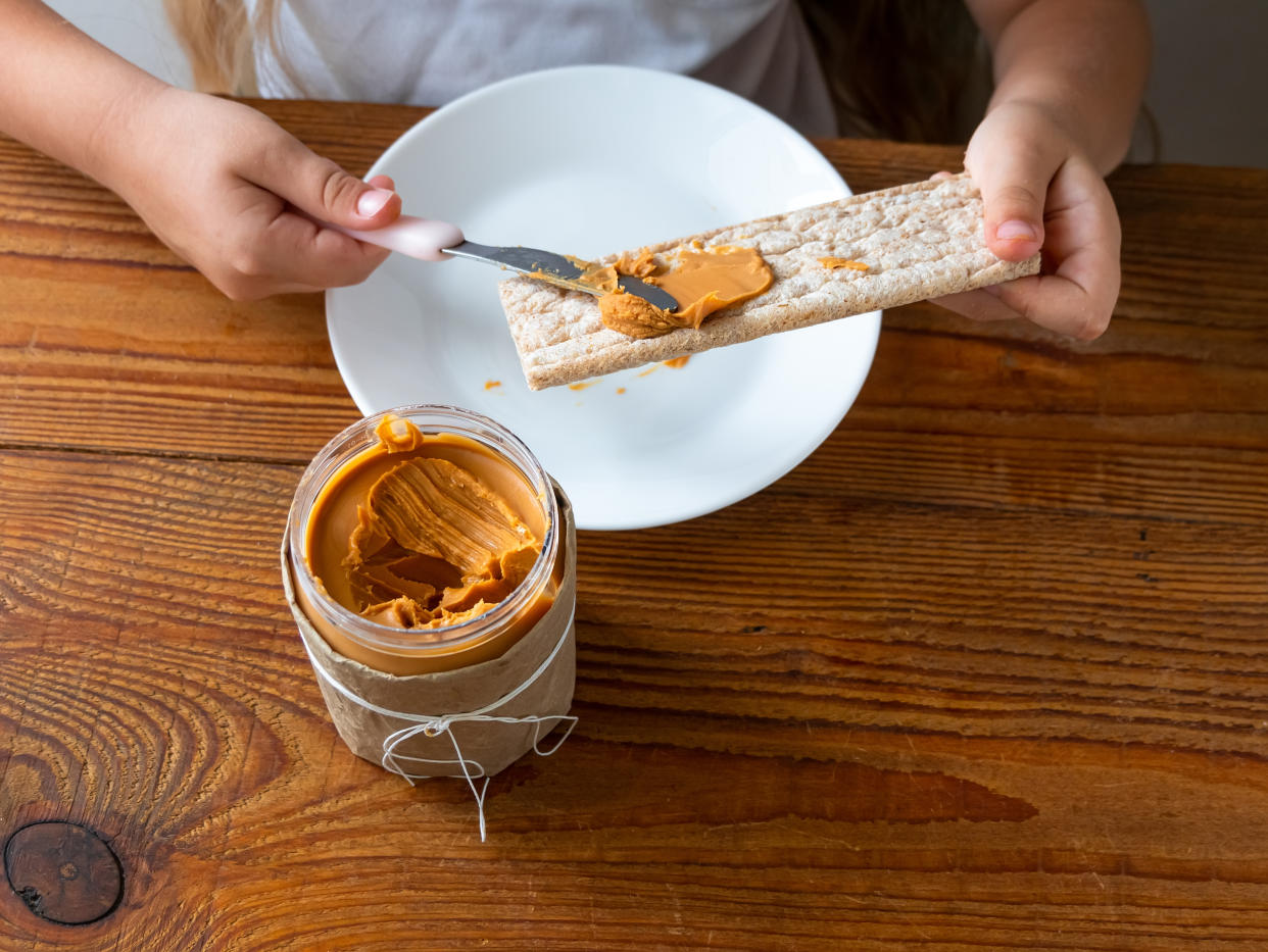 Child spreading peanut butter on crispbread eating sandwich wooden table home kitchen.School girl with bread slice wholegrain snack.Kid cooking breakfast Organic nutritious superfood healthy lifestyle�